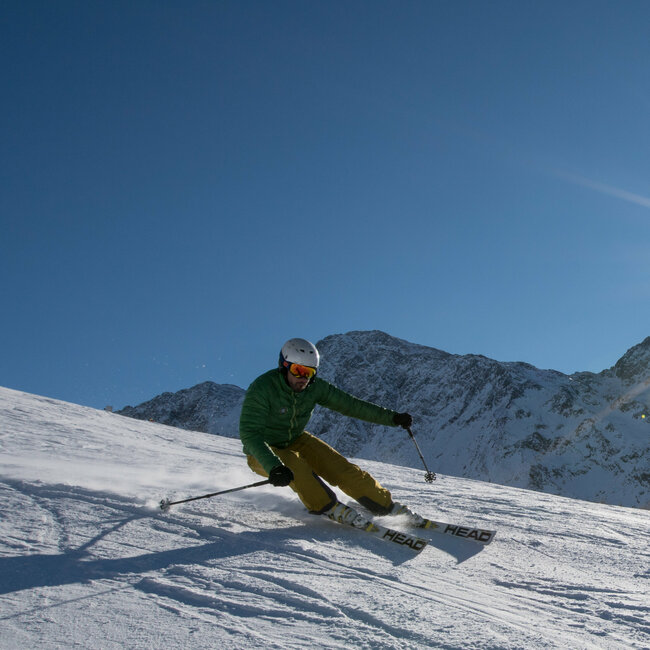 Ein Skifahrer mit grüner Jacke und gelber Hose carvt mit der Sonne im Rücken im Skigebiet Zettersfeld.