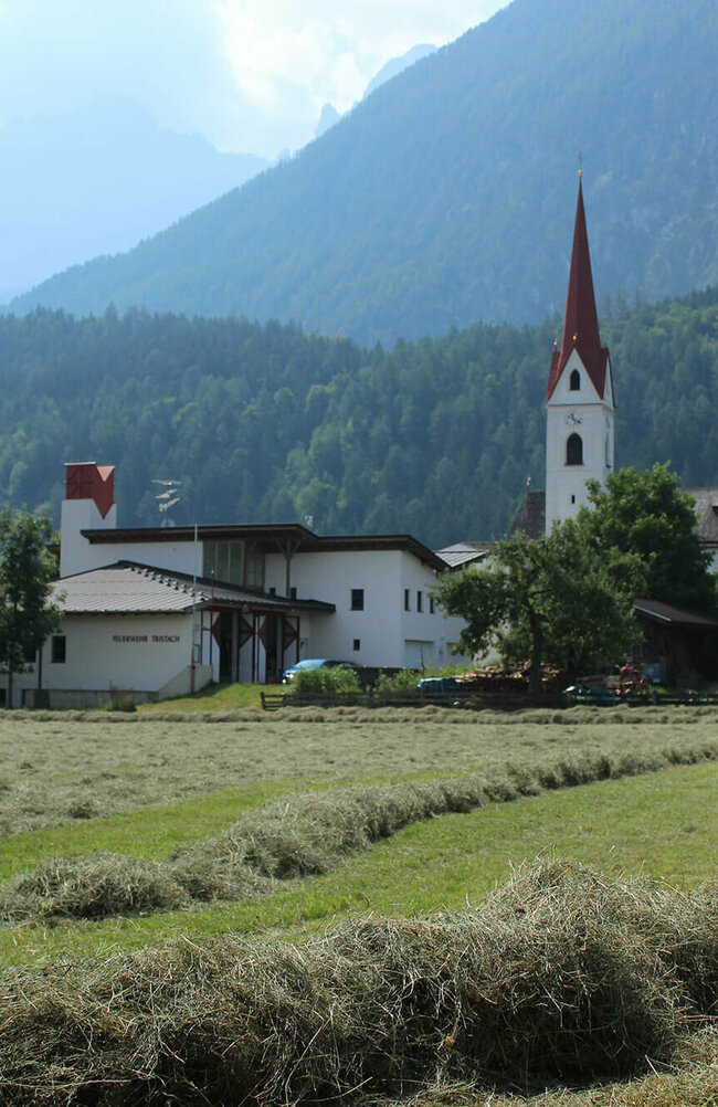 Tristach Die Tristacher Kirche, das Feuerwehr Gebäude und ein abgemähtes Feld mit liegendem Heu.