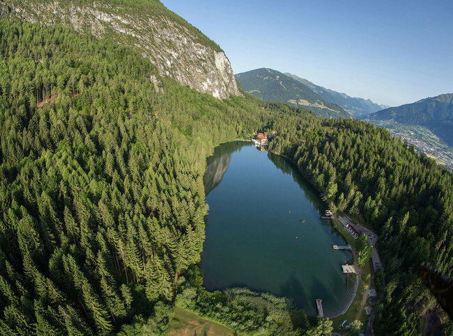 Tristacher See Luftaufnahme vom sanft in den Wald eingebetteten Tristacher See bei herrlichem Wetter, mit saftig grünen Bäumen drum herum.