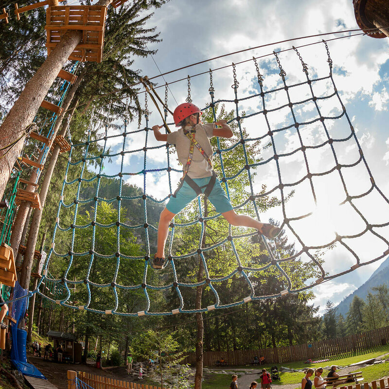 Hochseilgarten Galitzenklamm Kinder an einer Kletterstation im Hochseilgarten der Galitzenklamm bei sonnigem Wetter. Im Hintergrund spielen mehrere Personen am Wasserspielplatz.
