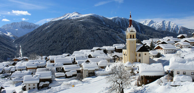 Obertilliach - zauberhaft verschneit im Winter Viel Neuschnee liegt auf den Dächern des Dorfes mit der prägnanten Kirche im Vordergrund an einem fast wolkenlosen Wintertag. Im Hintergrund bewaldete und verschneite Bergrücken sowie tief verschneite Landschaft oberhalb der Baumgrenze mit zum Teil bizarr geformtem Bergkamm.