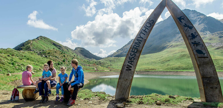 Wassermythos Ochsenlacke St. Jakob Brunnalm Eine Familie sitzt auf einer Bank am Ufer des Wassermythos Ochsenlacke im Skizentrum St. Jakob i. D.. Im Hintergrund sieht man die dazugehörigen Spielgeräte und das umliegende Bergpanorama, welches durch das schöne Wetter gut zur Geltung kommt.
