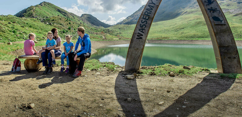 Wassermythos Ochsenlacke St. Jakob Brunnalm Eine Familie sitzt auf einer Bank am Ufer des Wassermythos Ochsenlacke im Skizentrum St. Jakob i. D.. Im Hintergrund sieht man die dazugehörigen Spielgeräte und das umliegende Bergpanorama, welches durch das schöne Wetter gut zur Geltung kommt.