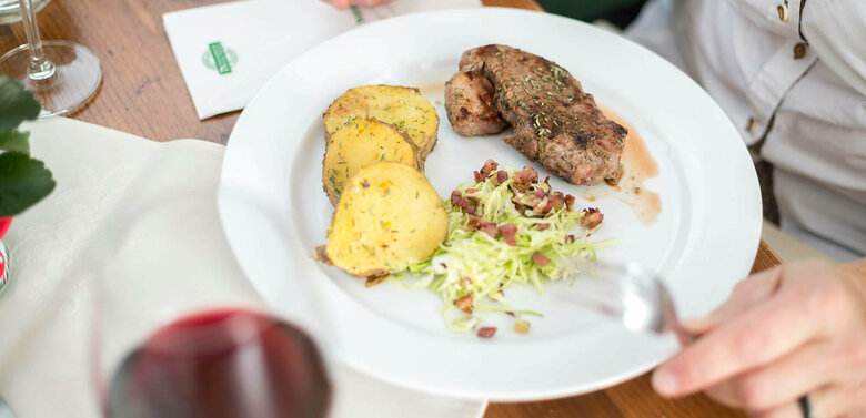 Regionsprodukte Gebratenes Fleischstück mit dicken Kartoffelscheiben und Krautsalat mit Speckwürfeln schön auf einem weißen Teller angerichtet mit einem Glas Rotwein.