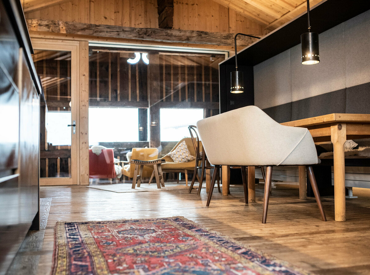 Innenansicht der Ferienwohnung Bergsteiger SEIN im Kesslerstadel aus der Ameisenperspektive mit einem gemusterten Teppich auf dem Holzboden, einem Esstisch mit gepolsterter Bank und Wintergarten mit gemütlichen Stühlen.