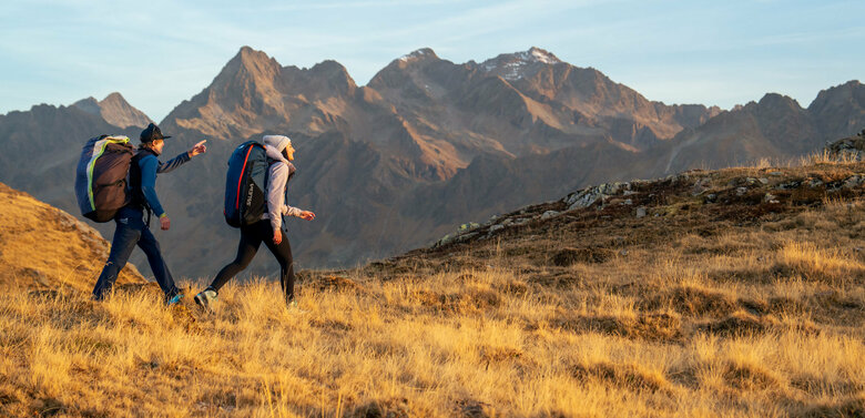 Ein Mann und eine Frau mit großen Gleitschirm-Rucksäcken auf dem Rücken gehen auf einer herbstlichen Wiese auf dem Zettersfeld.
