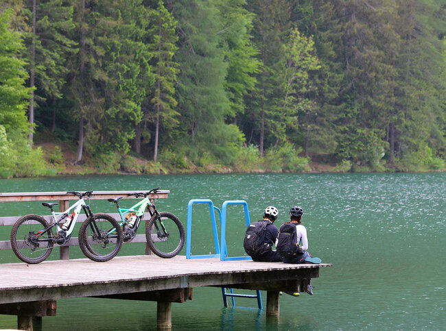 E Biken Ein Pärchen sitzt auf dem Holzsteg am Tristacher See und blickt aufs Wasser. Hinter ihenen stehen ihre E-Bikes.