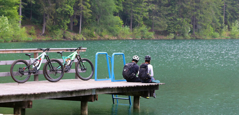 E Biken Ein Pärchen sitzt auf dem Holzsteg am Tristacher See und blickt aufs Wasser. Hinter ihenen stehen ihre E-Bikes.