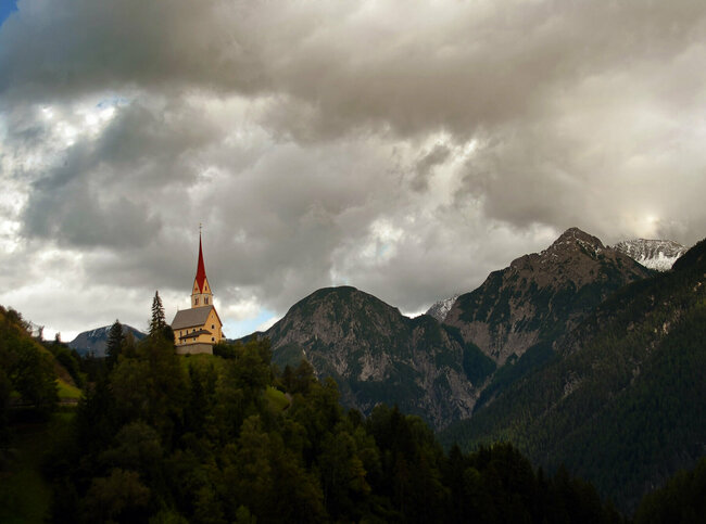 Kirche St. Justina Kirche St. Justina inmitten eines Hanges umgeben von Bäumen und den Lienzer Dolomiten im Hintergrund.