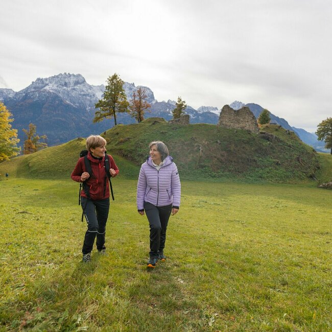 Wandern Ruine Walchenstein Zwei Damen wandern über eine grüne Wiese und lächeln sich zu. Im Hintergrund sieht man die Ruine Walchenstein.