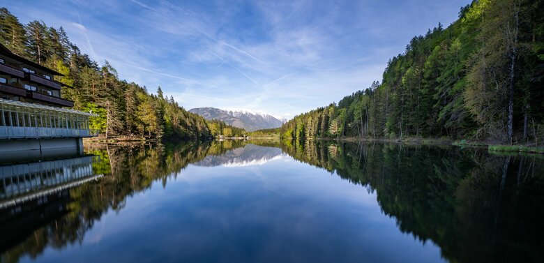 Blick vom Hotel am Tristachersee auf den glasklaren See. Die Bäume spiegeln sich am Rande im Wasser.