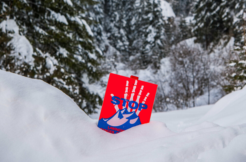 Ein rot-weiß-blaues Warnschild steckt im tiefen Schnee und warnt vor Lawinen.
