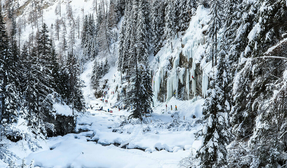Eiskletterpark Osttirol Drei Eiskletterer stehen vor der Eiswand und sehen auf ihr nach oben. Umrahmt wird der Eiskletterpark von tief verschneiten dunkelgrünen Tannenbäumen.