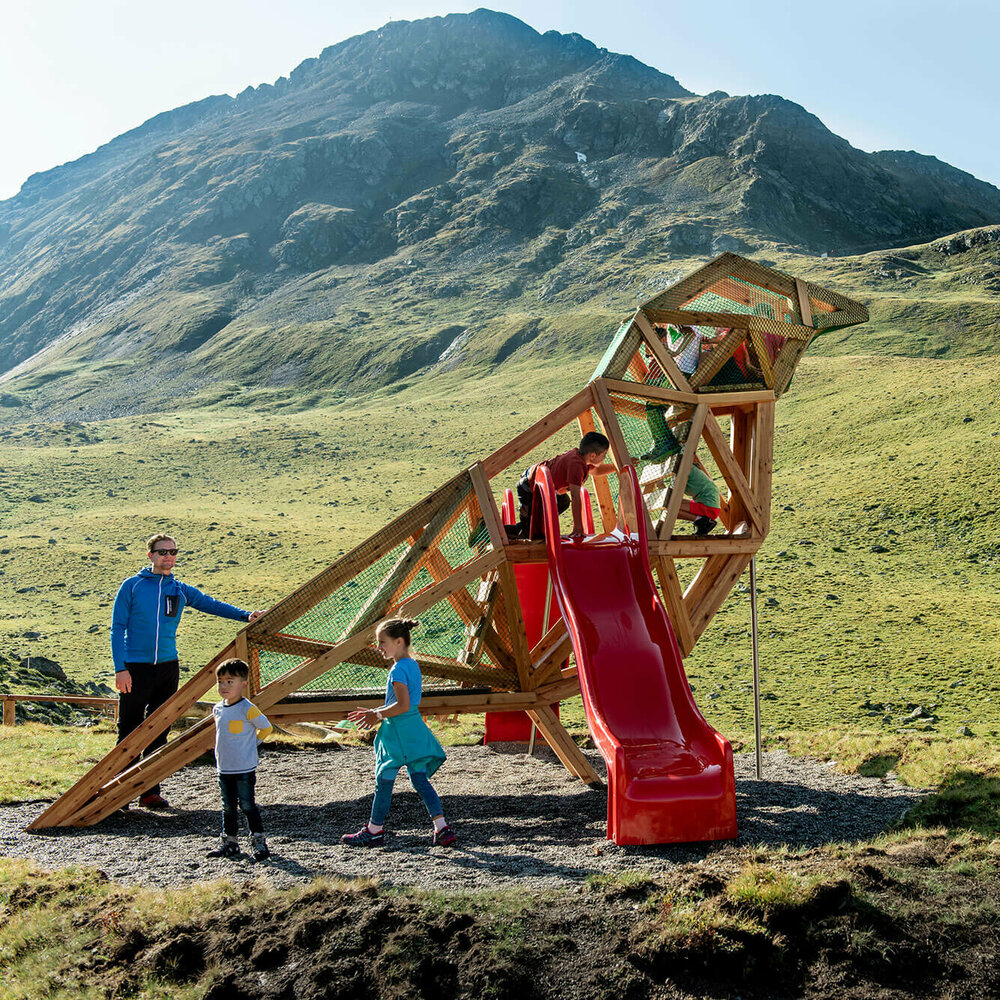 Wassermythos Ochsenlacke St. Jakob Brunnalm Zu sehen sind glückliche Kinder auf einem Spielgerät, welches einem überdimensional-großen Vogel ähnlich sieht, des Wassermythos Ochsenlacke im Skizentrum St. Jakob i. D.. Das sonnige Wetter lässt die umliegende Umgebung und die Berge im Hintergrund in einem warmen Licht erstrahlen.