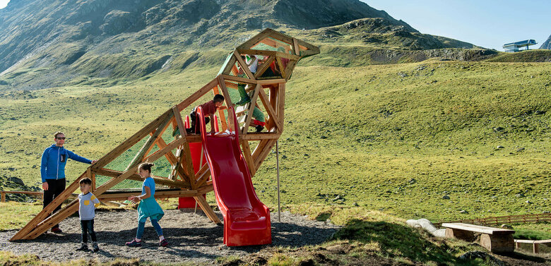 Wassermythos Ochsenlacke St. Jakob Brunnalm Zu sehen sind glückliche Kinder auf einem Spielgerät, welches einem überdimensional-großen Vogel ähnlich sieht, des Wassermythos Ochsenlacke im Skizentrum St. Jakob i. D.. Das sonnige Wetter lässt die umliegende Umgebung und die Berge im Hintergrund in einem warmen Licht erstrahlen.
