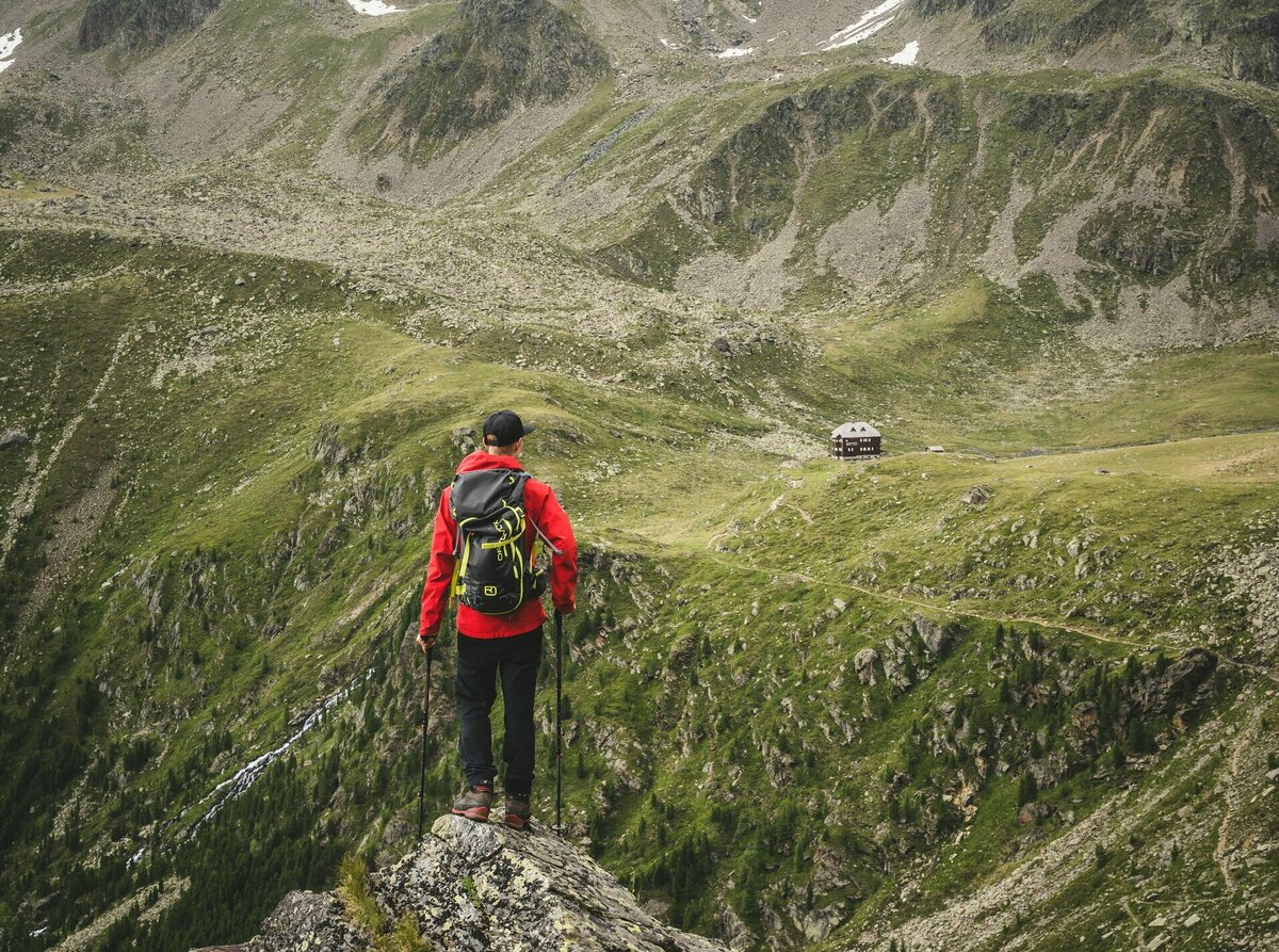 Hochschoberhütte Ein Wanderer mit roter Jacke und Rucksack steht auf einem Felsen mit Blick auf die Hochschoberhütte in Osttirol.