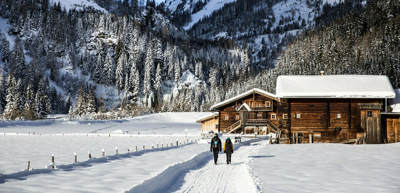 Zwei Wanderer gehen nebeneinander, inmitten tief verschneiter Winterlandschaft, auf einem Winterwanderweg. Im Hintergrund sieht man zwei dunkelbraune, schneebedeckte Almhütten und einen tief grünen Nadelbaumwald mit verschneiten Spitzen.