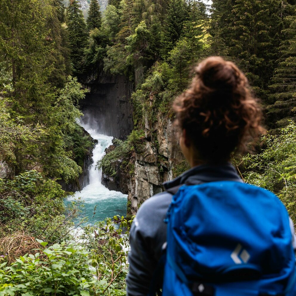 Frau mit Wasserfall Eine Frau mit blauem Rucksack und braunem Dutt steht vor einem Wasserfall mit Blickrichtung zum Wasser.