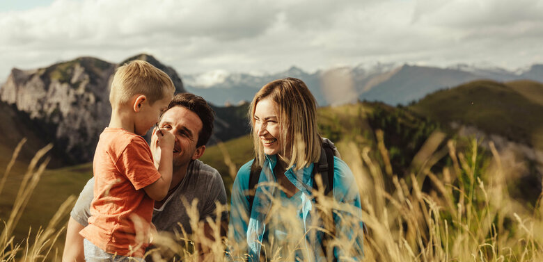 kindermeile-obertilliach Zu sehen ist ein Elternpaar mit ihrem kleinen Sohn, die im hohen Gras entlang der Kindermeile am Golzentipp den Moment zu dritt genießen.