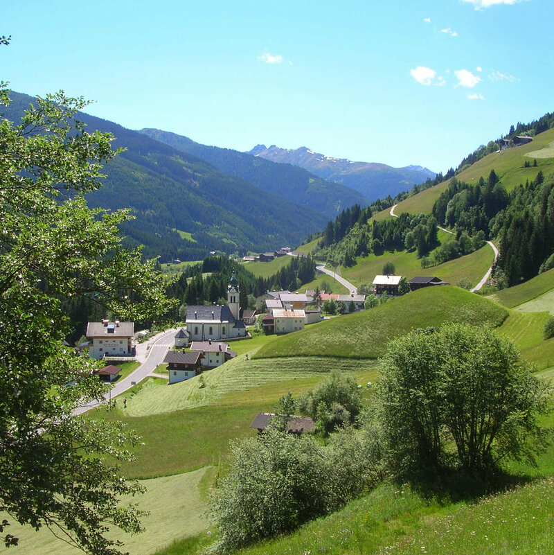 Untertilliach Blick von einem höher gelegenen Hang auf die Pfarrkirche in Untertilliach. Auf den umliegenden saftig grünen Wiesen finden Mäharbeiten bei strahlend blauem Himmel statt.
