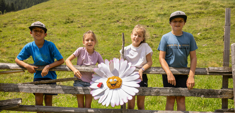 Die vier Kinder stehen mit Gisela Gänseblümchen in ihrer Mitte auf einem Holzzaun vor einer grünen Wiese.