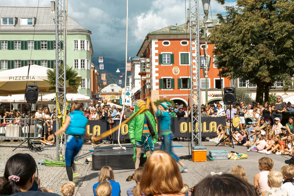 Drei Artistinnen am Olala 2025, zwei davon hüllen die dritte in dicke Winterkleidung, rundherum stehen und sitzen begeisterte Zuseherinnen und Zuseher.