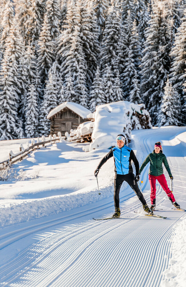 Zwei Langläufern:innen auf der Langlaufloipe in Obertilliach mit einem frisch verschneitem Wald im Hintergrund