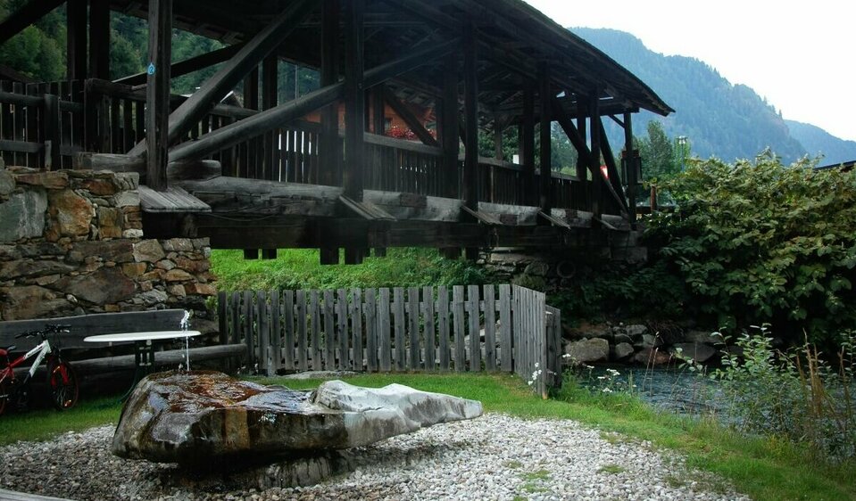 Wasserruheplatz Blosbrücke Der Wasserruheplatz bei der Blosbrücke in Hopfgarten in Defereggen in den Sommermonaten. Das grüne Gras bedeckt das Ufer neben der blauen Schwarzach, über welche die robuste Holzbrücke ragt.