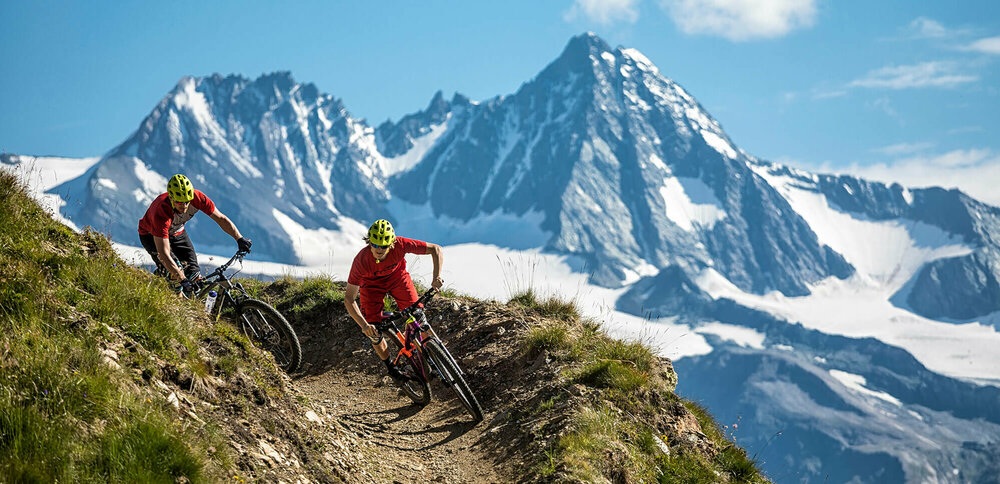 Bikepark Grossglockner Resort Zwei Radfahrer mit roten Shirts und gelben Helmen auf einem schmalen Trail im Bikepark Grossglockner Resort. Im Hintergrund ragt der Großglockner in den blauen Himmel.