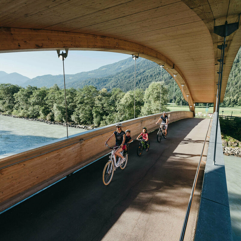 Drauradweg Die Abbildung zeit eine Familie, die mit ihren Fahrrädern auf dem Drauradweg unterwegs sind, und gerade eine hölzerne Brücke überqueren.