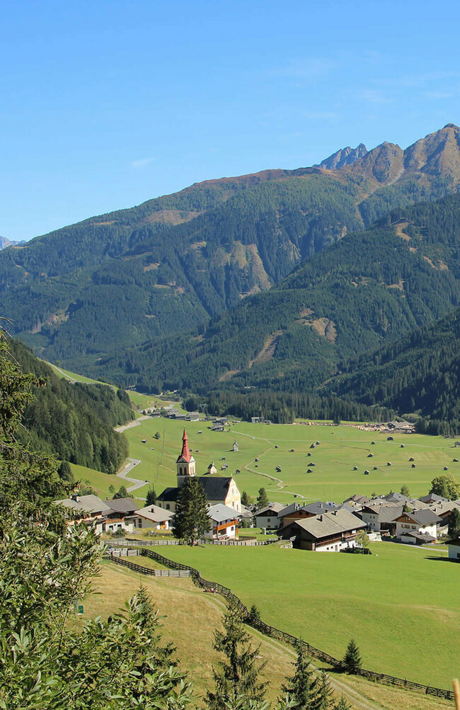Obertilliach Blick auf eng verbautes Haufendorf mit ausschließlich grauer Dacheindeckung an einem wolkenlosen Herbsttag. Im Hintergrund die grünen Felder mit vielen kleinen Heuhütten und ein Gebirgszug, der das Tal mit seinen bewaldeten Hängen verschließt.
