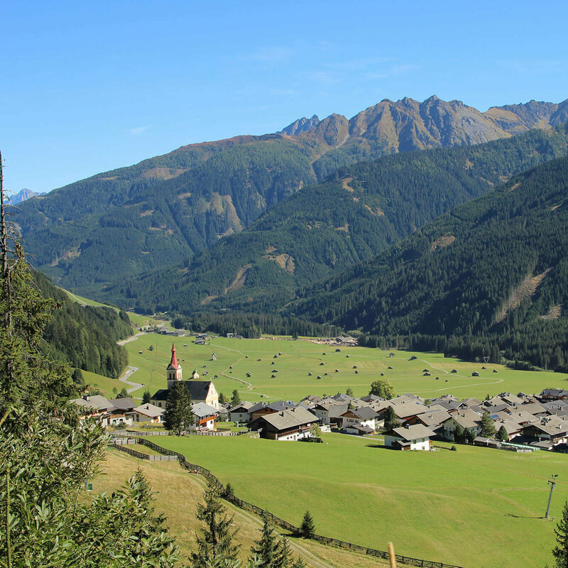 Obertilliach Blick auf eng verbautes Haufendorf mit ausschließlich grauer Dacheindeckung an einem wolkenlosen Herbsttag. Im Hintergrund die grünen Felder mit vielen kleinen Heuhütten und ein Gebirgszug, der das Tal mit seinen bewaldeten Hängen verschließt.