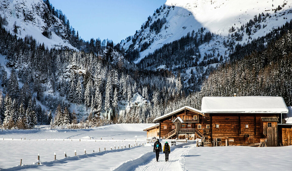 Eiskletterpark Osttirol Zwei Personen spazieren auf einem Winterwanderweg an einer kleinen Ansammlung von kleinen Hütten vorbei. Dahinter der bewaldete Talschluss in einer winterlichen Umgebung.