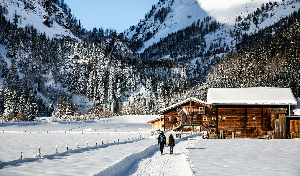 Eiskletterpark Osttirol Zwei Personen spazieren auf einem Winterwanderweg an einer kleinen Ansammlung von kleinen Hütten vorbei. Dahinter der bewaldete Talschluss in einer winterlichen Umgebung.
