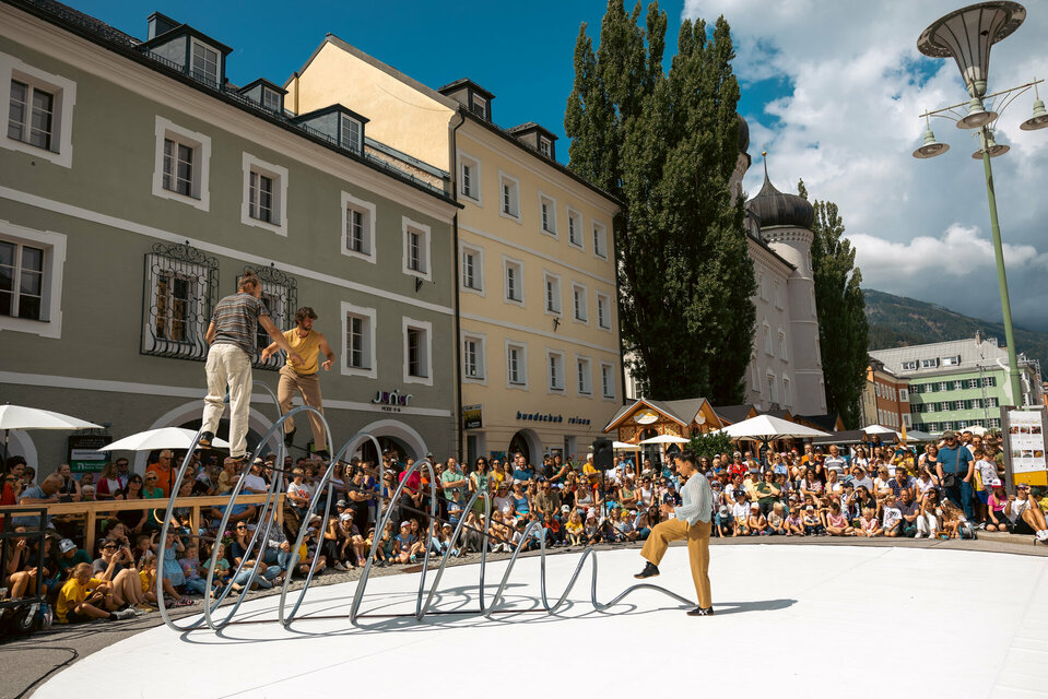 Drei Artisten turnen auf, in bzw. vor einer großen Spirale aus Eisen am Lienzer Hauptplatz im Rahmen von Olala 2025. Sie werden vom begeisterten Publikum rundherum bestaunt.