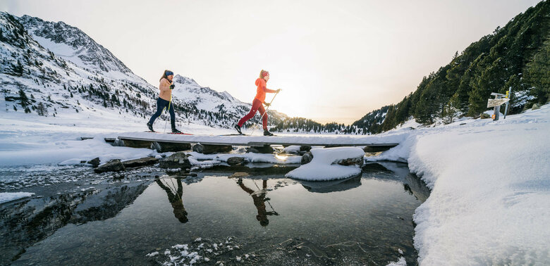 zwei Langläufer:innen überqueren einen Bach beim Langlaufen im Defereggental