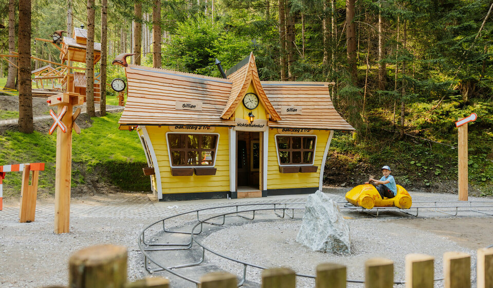 Auf dem Bild ist der neue Bahnhof des Wichteldorfes zu sehen, vor dem ein kleiner Junge mit einem Miniatur Tretauto vor dem Bahnhof seine Runden dreht.
