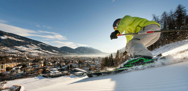 Skifahren Hochstein Ein Skifahrer mit gelber Jacke und heller Hose carved am Hochstein mit den Ski. Aufgenommen von unten. Blick auf das Dorf.