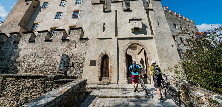 Biken Schloss Bruck Zwei Biker schieben ihre Räder durch einen steinernen Bogen am Eingang des Schloss Bruck in Lienz.