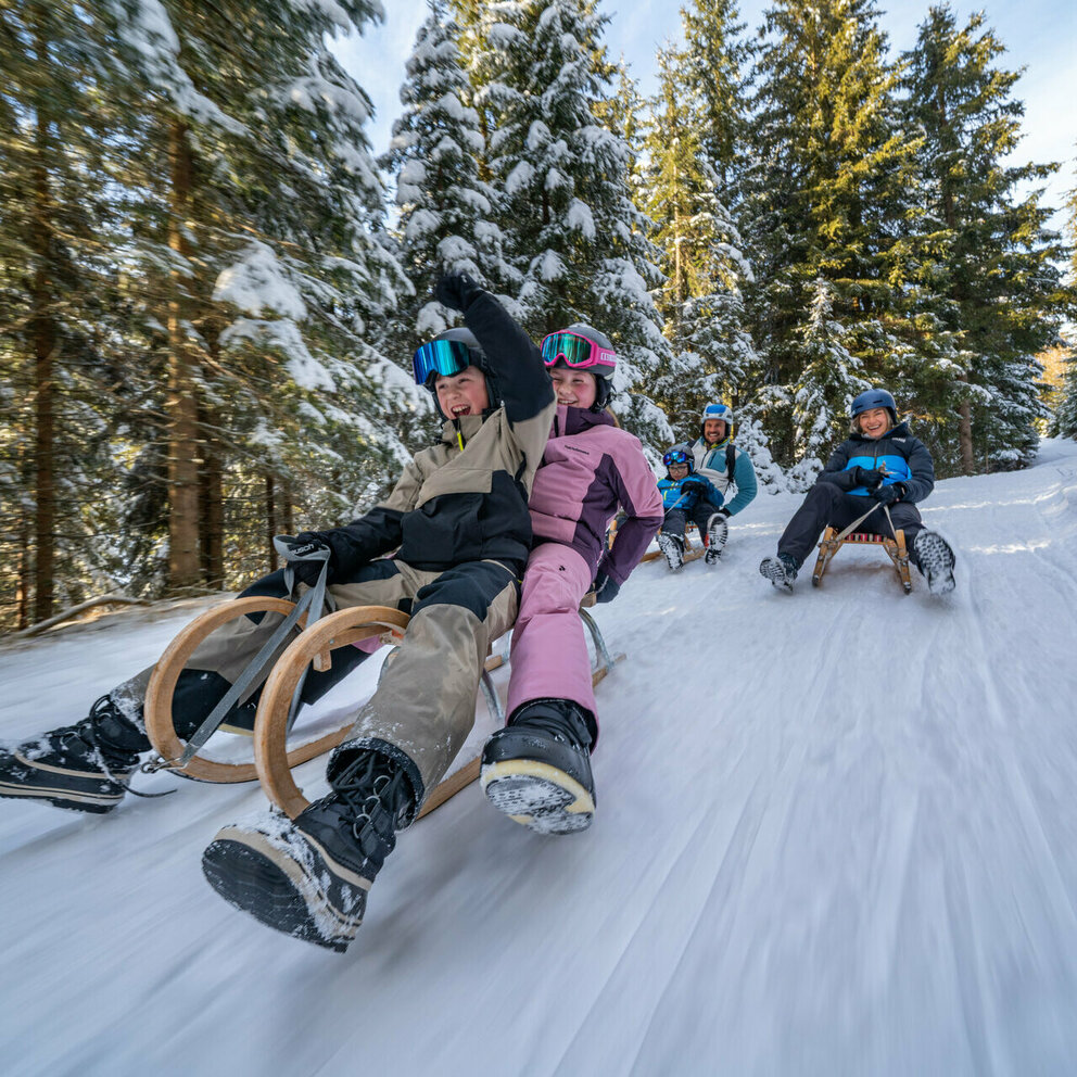 Eine fünfköpfige Familie in bunten Skianzügen auf der rasanten Rodelabfahrt in Außervillgraten.