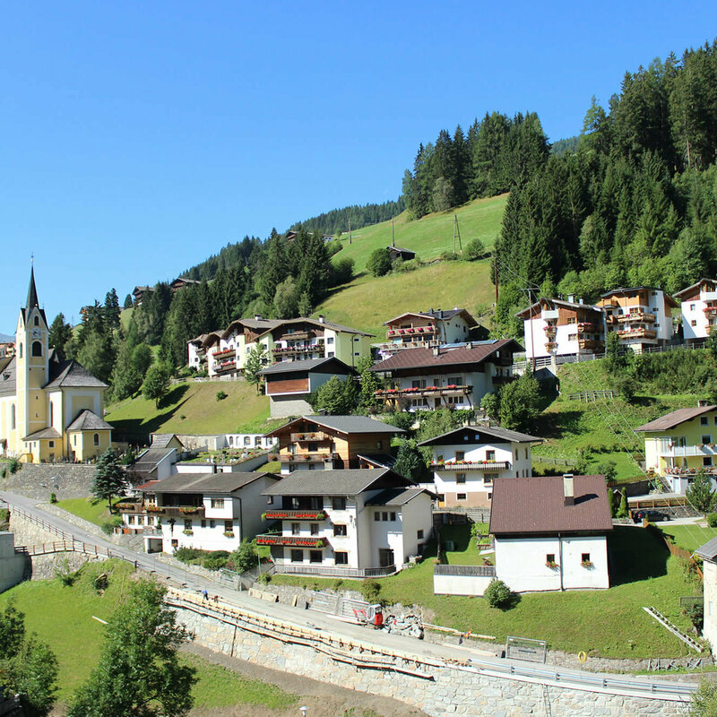 Ausservillgraten Das Bild zeigt das Dorf Außervillgraten mit seiner Kirche und einer Wohnsiedlung auf saftig grünem Untergrund, gelegen unter strahlend blauem Himmel.