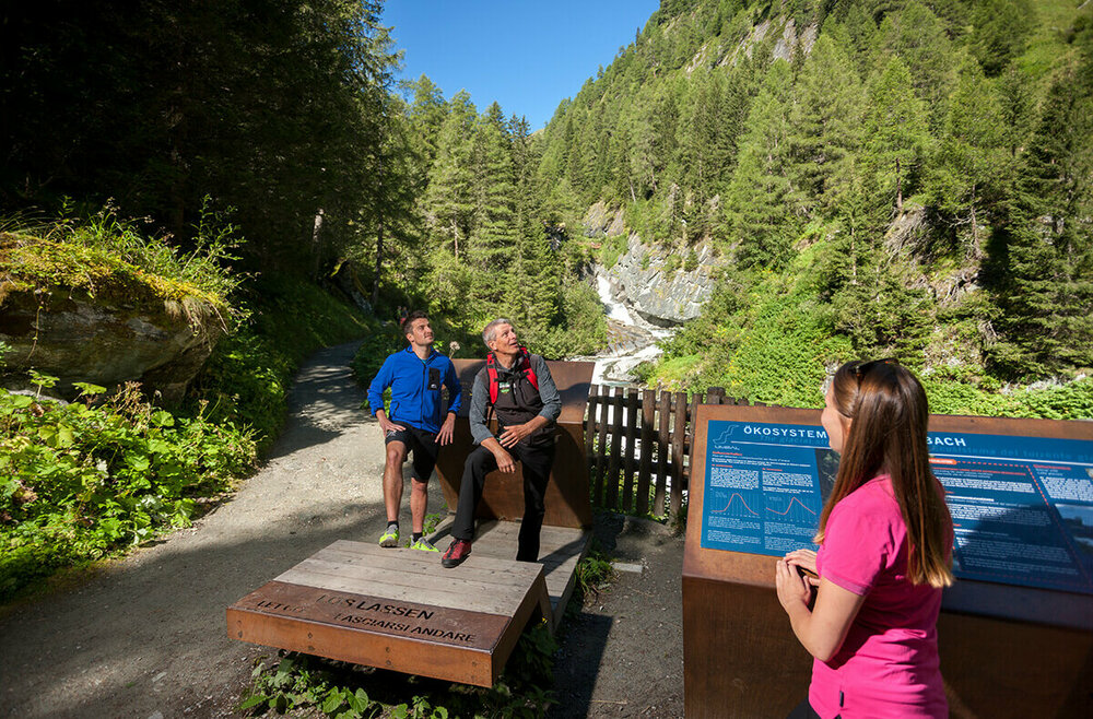 Umballfälle Lehrpfad An einem Sommertag im Nationalpark Hohe Tauern betrachten drei Personen eine Informationstafel, während im Hintergrund die Isel als Gletscherfluss durch die Landschaft fließt