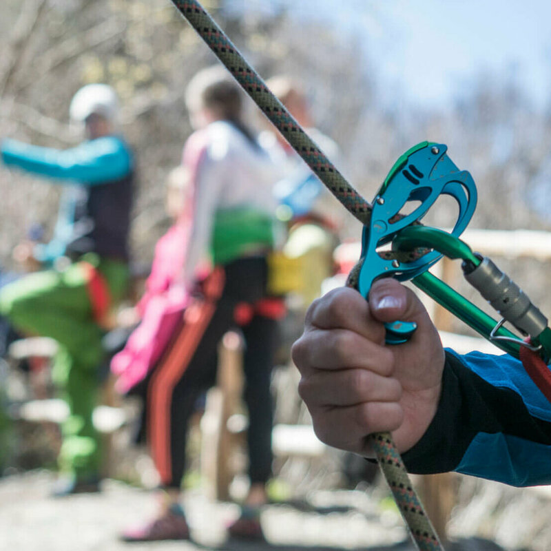 Karabiner Nahaufnahme von einem Kletter-Sicherungsgerät, einem Seil und bunten Karabinern. Im Hintergrund stehen mehrere Personen.
