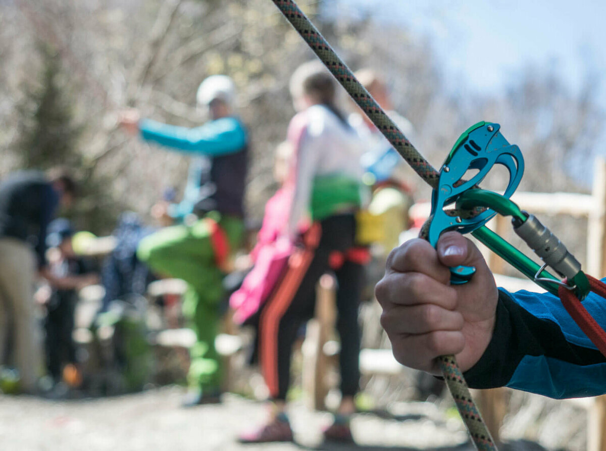 Karabiner Nahaufnahme von einem Kletter-Sicherungsgerät, einem Seil und bunten Karabinern. Im Hintergrund stehen mehrere Personen.