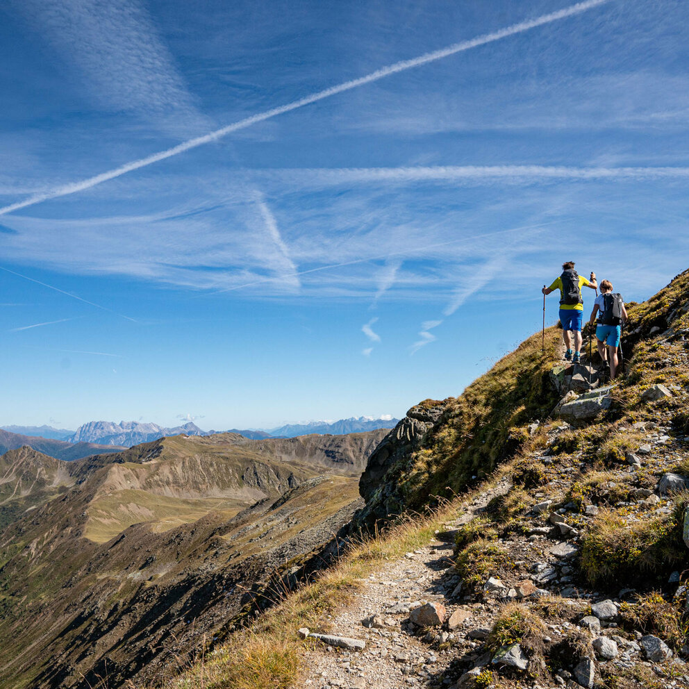 Herz-Ass Villgratental Das Bild zeigt zwei Wanderer auf der Strecke der Herz-Ass-Villgratental Wanderung mit imposanten Blick über die entfernten Berggipfel, die unter strahlend blauen Himmel ruhen.