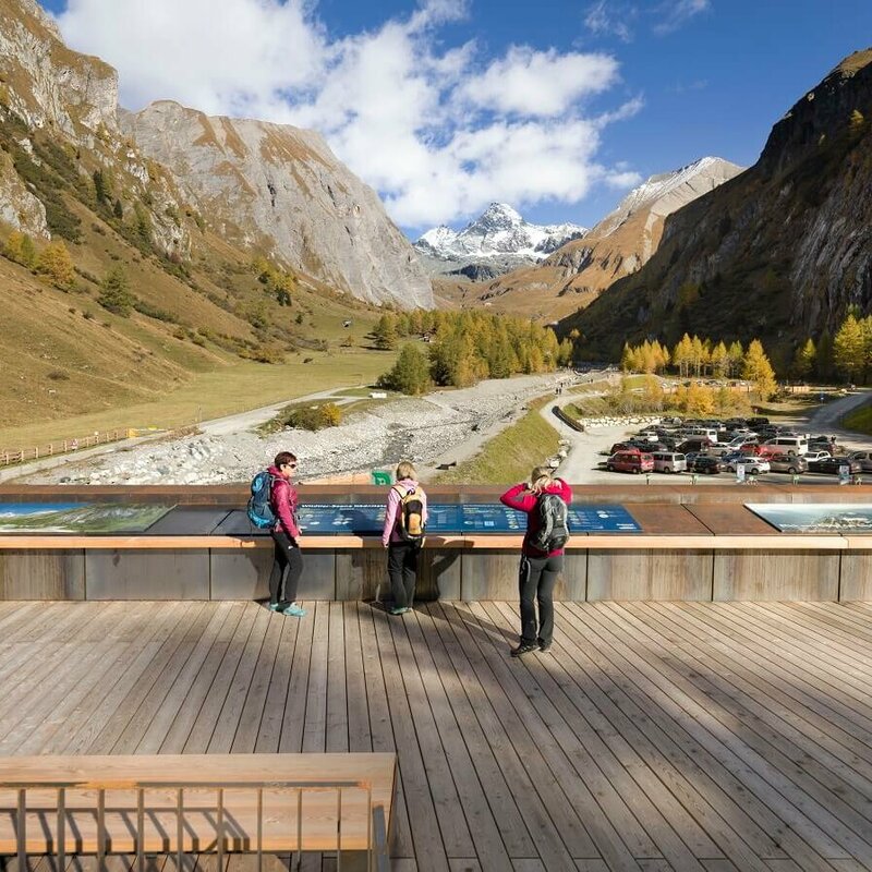 Kalser Glocknerstraße Drei Frauen stehen auf einem Parkplatz beim Glocknerwinkel in Kals und blicken auf den Großglockner. Die herbstliche Landschaft zeigt bunte Bäume und klare Sicht auf den schneebedeckten Berg.