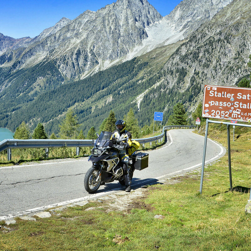 Motorradfahrer am Staller Sattel. Neben ihm befindet sich ein Verkehrsschild mit der Aufschrift Staller Sattel. Im Hintergrund befinden sich Berge.