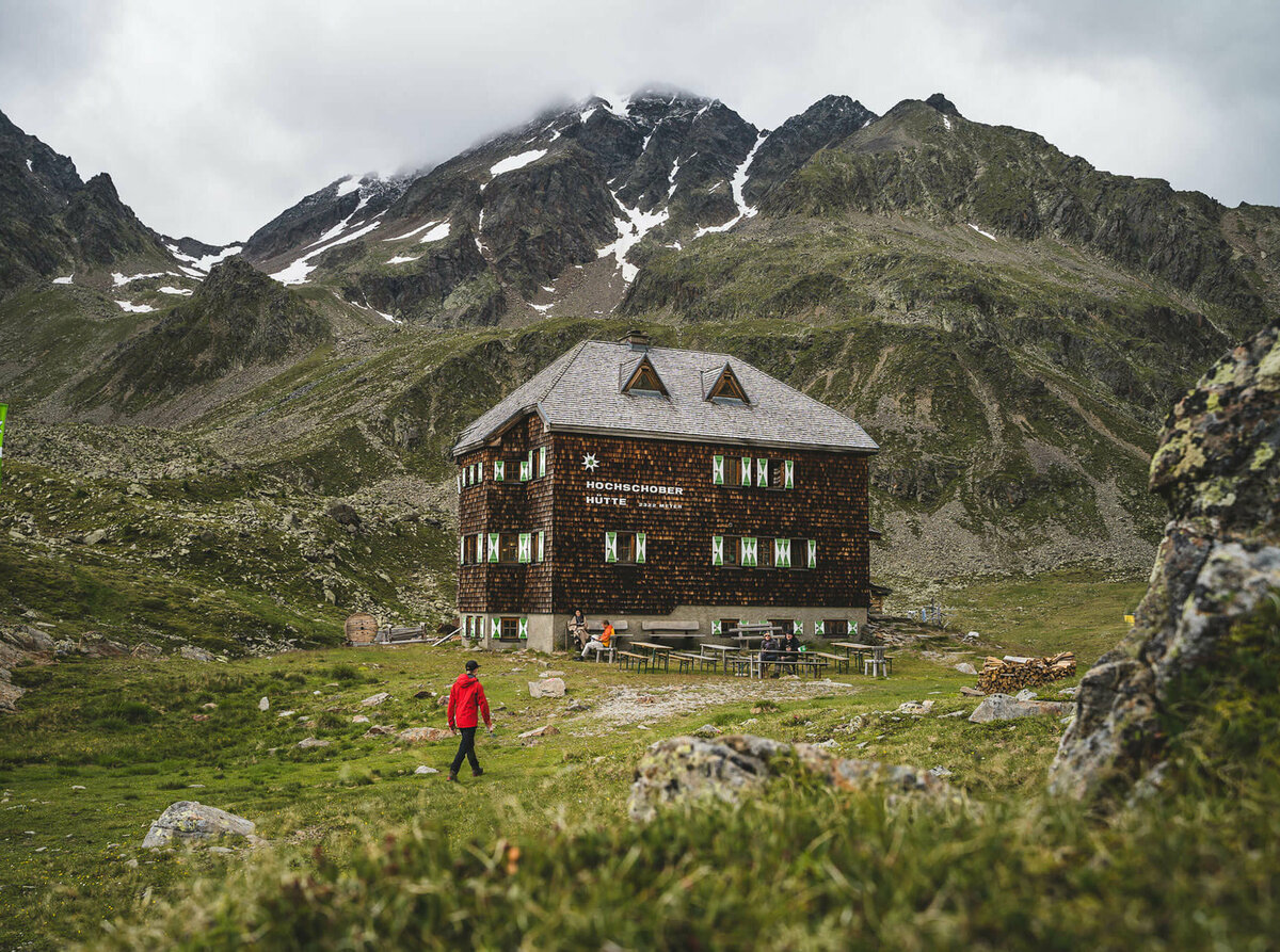 Hochschober Hütte Eine Person mit roter Jacke geht kurz vor der Hochschober Hütte. Links steht eine grüne Alpenvereins-Flagge.