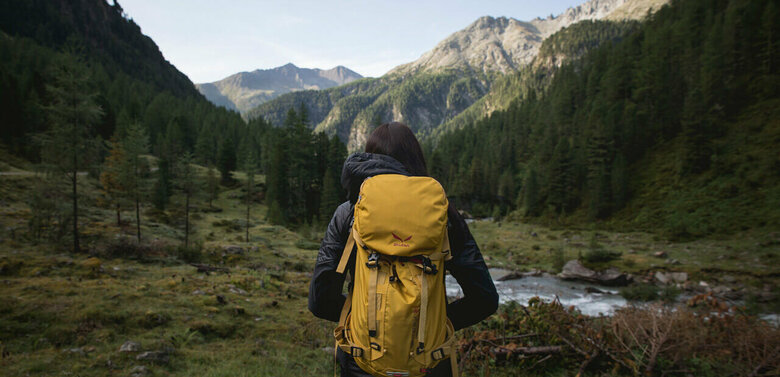 Wandern in Osttirol Wanderin mit gelbem Rucksack auf einer Lichtung in morgendlicher Stimmung kurz vor einem kleinen Gebirgsbach. Im Hintergrund strahlt die Morgensonne schon einen Bergrücken an.
