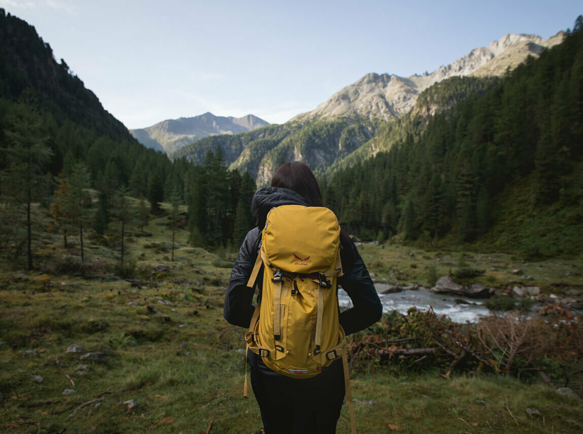 Wandern in Osttirol Wanderin mit gelbem Rucksack auf einer Lichtung in morgendlicher Stimmung kurz vor einem kleinen Gebirgsbach. Im Hintergrund strahlt die Morgensonne schon einen Bergrücken an.